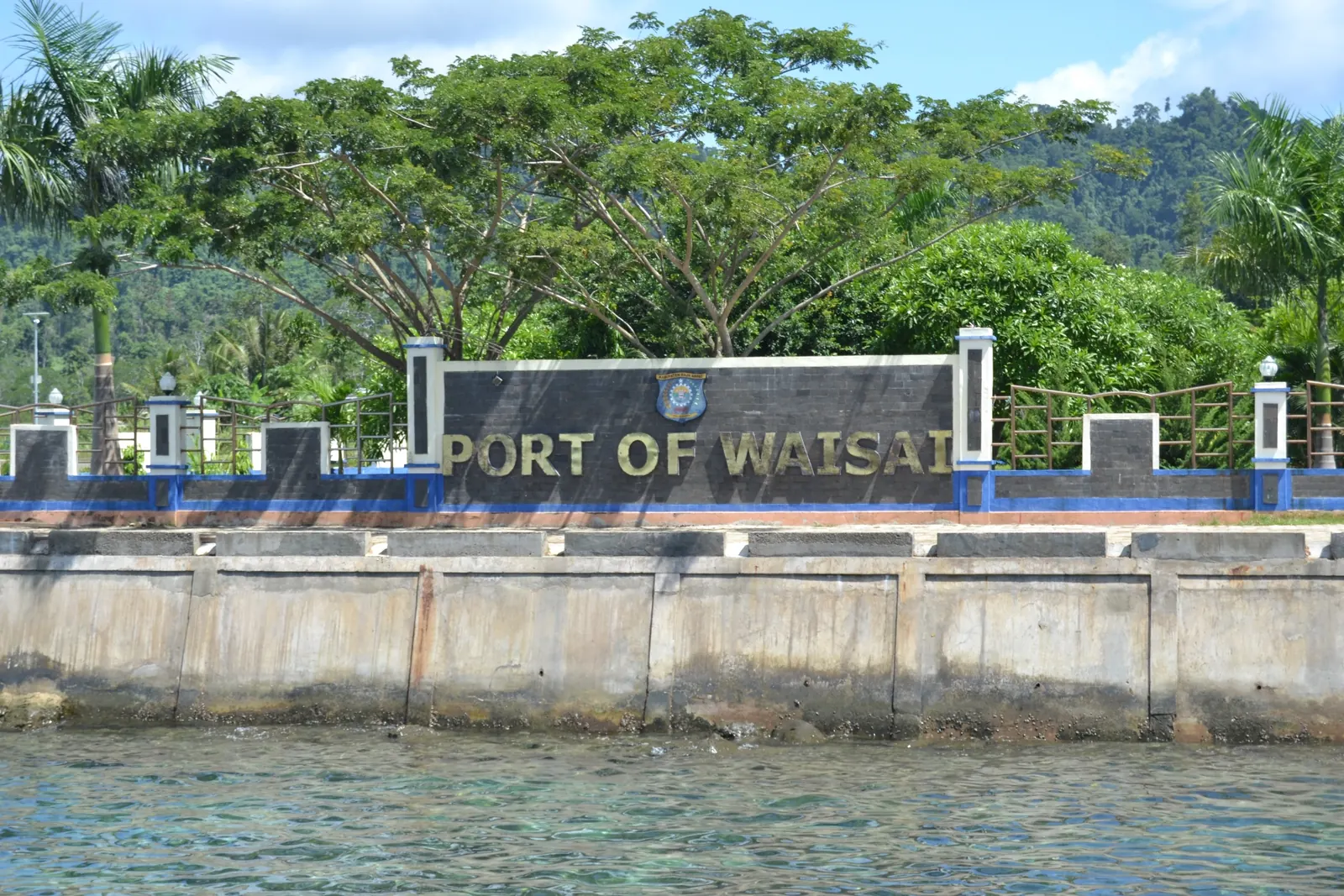 Tembok tepian Pelabuhan Waisai bertuliskan Port of Waisai, dengan pepohonan di belakang dan laut di depan