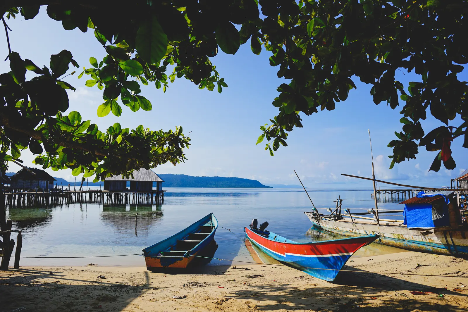 Perahu kayu berwarna biru dan merah di pantai Sawinggrai dengan bangunan panggung dan titian di atas air